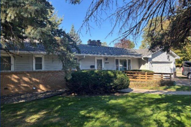 A single-story ranch-style house with a light exterior sits in a sunny front yard with green grass, shrubs, and a driveway on the right.
