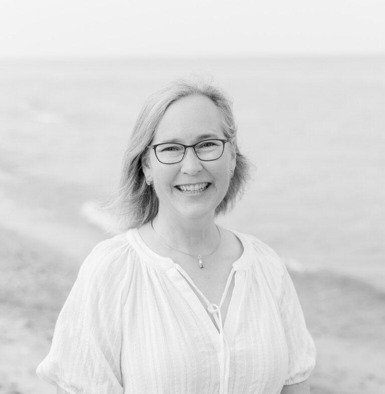Smiling woman with shoulder-length hair and glasses standing on a beach.