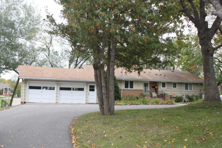 A single-story suburban home with an attached garage and a curved driveway, framed by large trees and a green front lawn.