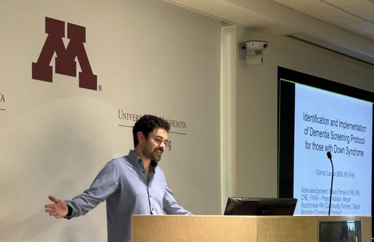 A man giving a lecture at a podium in a classroom, with a projected slide and a large maroon “M” logo on the wall.