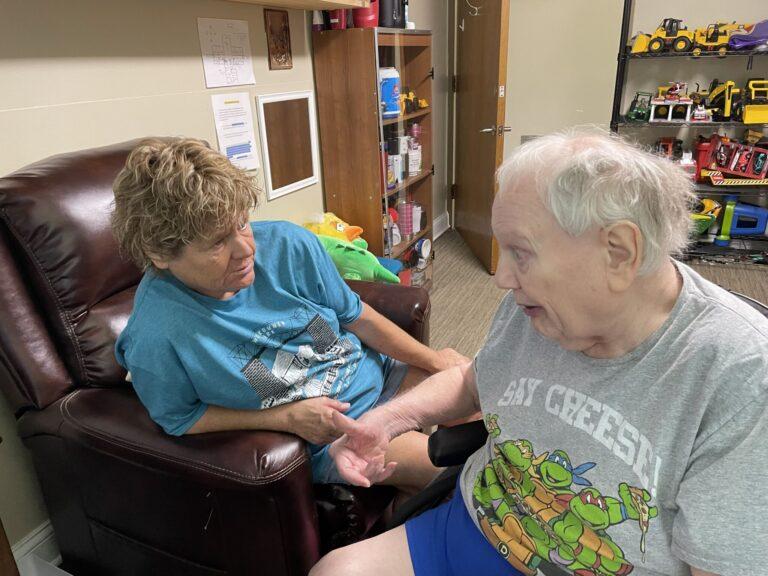 Two people sit facing each other and hold hands in a cozy room with a recliner and toy-filled shelves.