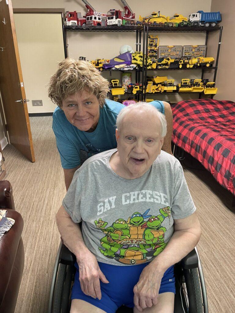 An elderly man in a wheelchair sits with a smiling caregiver behind him in a room filled with toy trucks on shelves.