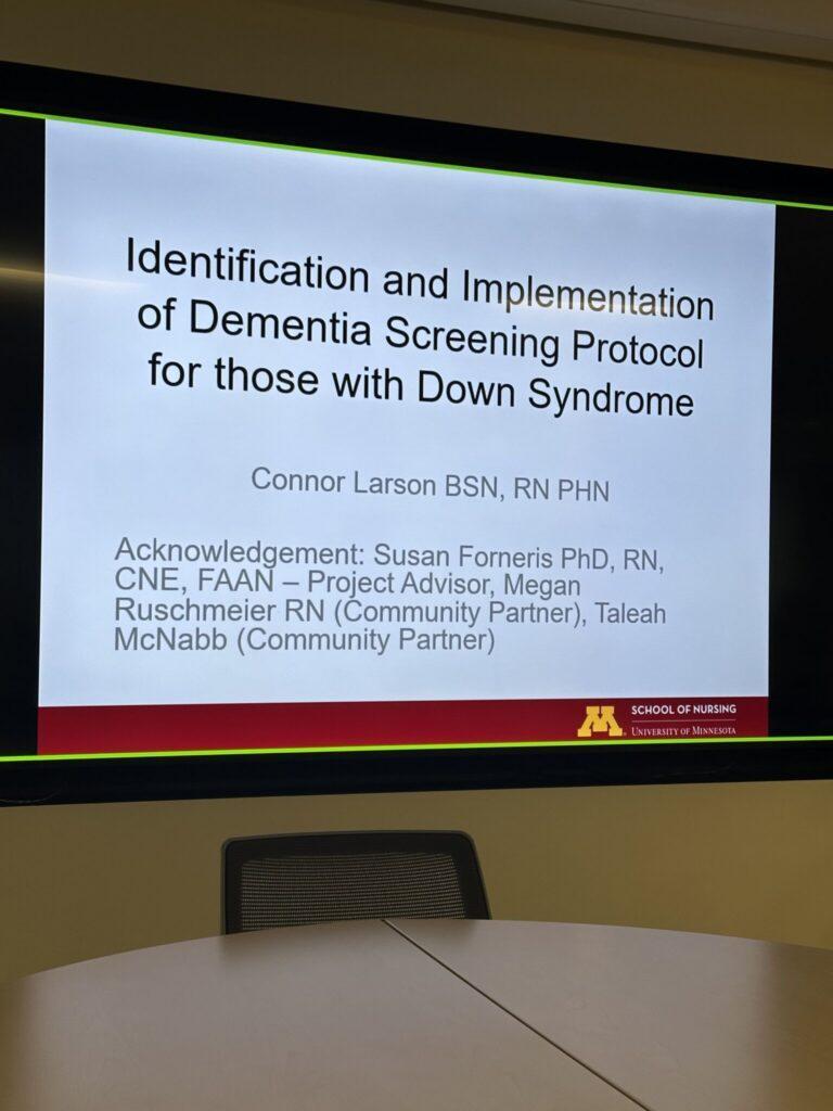 Presentation slide titled “Identification and Implementation of Dementia Screening Protocol for those with Down Syndrome” displayed in a conference room.
