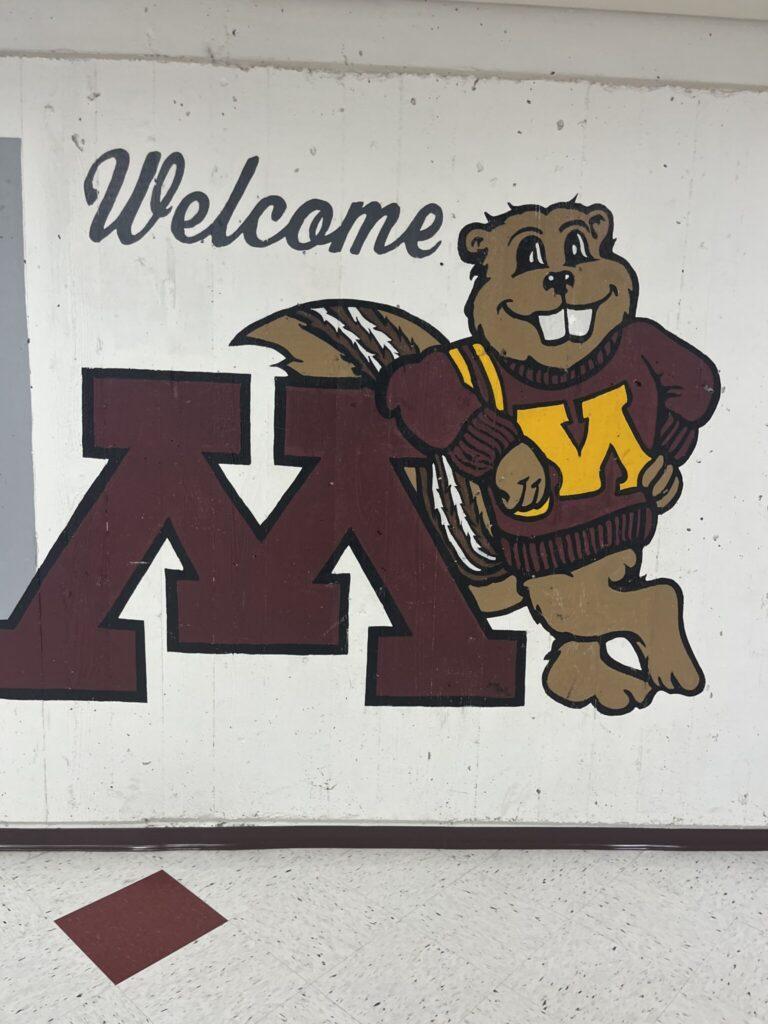 Cartoon bear mascot wearing a maroon and yellow shirt stands beside a large maroon "M" with a "Welcome" sign above.