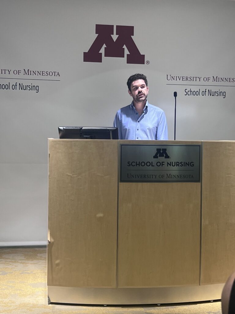 Man in a blue shirt stands at a wooden podium in front of a University of Minnesota School of Nursing backdrop featuring a large purple M logo.