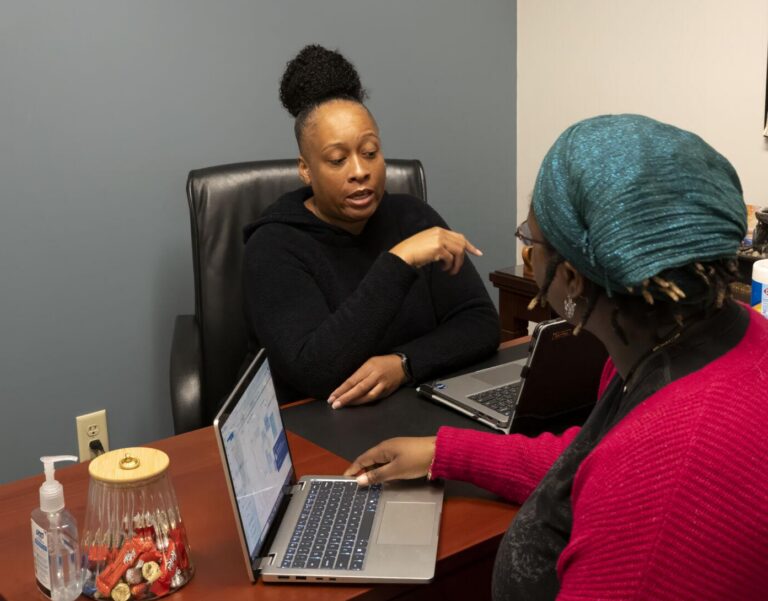 Two women sit across from each other at a desk in an office, one with a bun wearing black and the other wearing a teal headscarf and pink sweater, with laptops and office supplies on the desk.