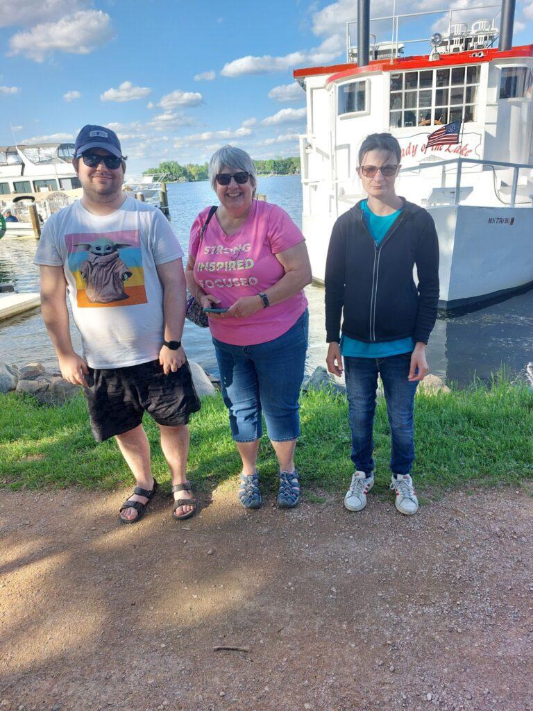 Three people stand on a dirt path in front of a docked boat at the marina, posing for a photo.