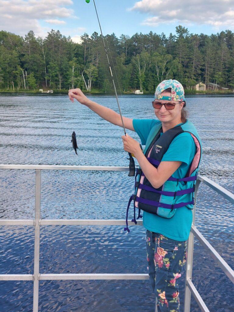 Person on a boat wearing a blue life jacket holds a fishing line with a small fish on the hook, with a lake and trees in the background.