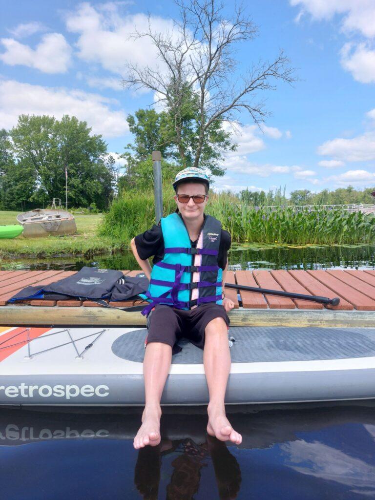 Person wearing a blue life jacket sits on a paddleboard by a wooden dock on a calm lake.