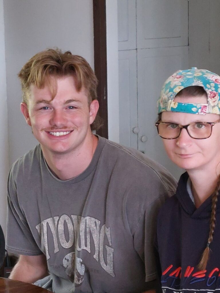 Two young adults sit indoors side by side: a smiling blond man in a gray T-shirt on the left and a woman with glasses, braided hair, and a colorful cap worn backward on the right.