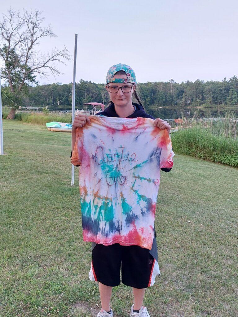 A person wearing a blue cap and glasses holds up a colorful tie-dye cloth outdoors on a grassy field.