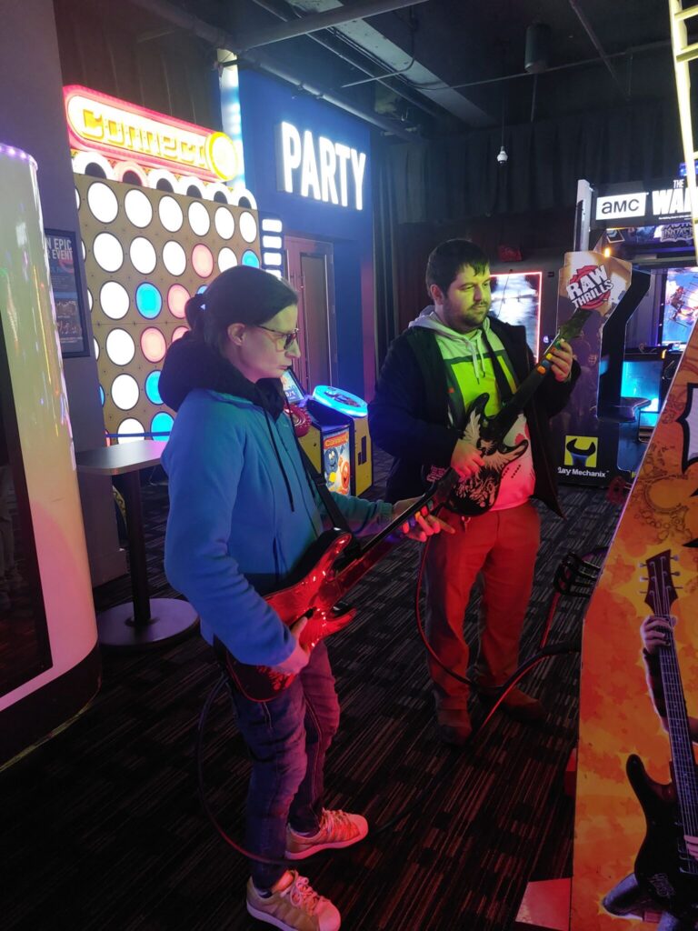 Two players in a colorful arcade, one holding a red toy gun, with neon signs including a large "PARTY" sign in the background.