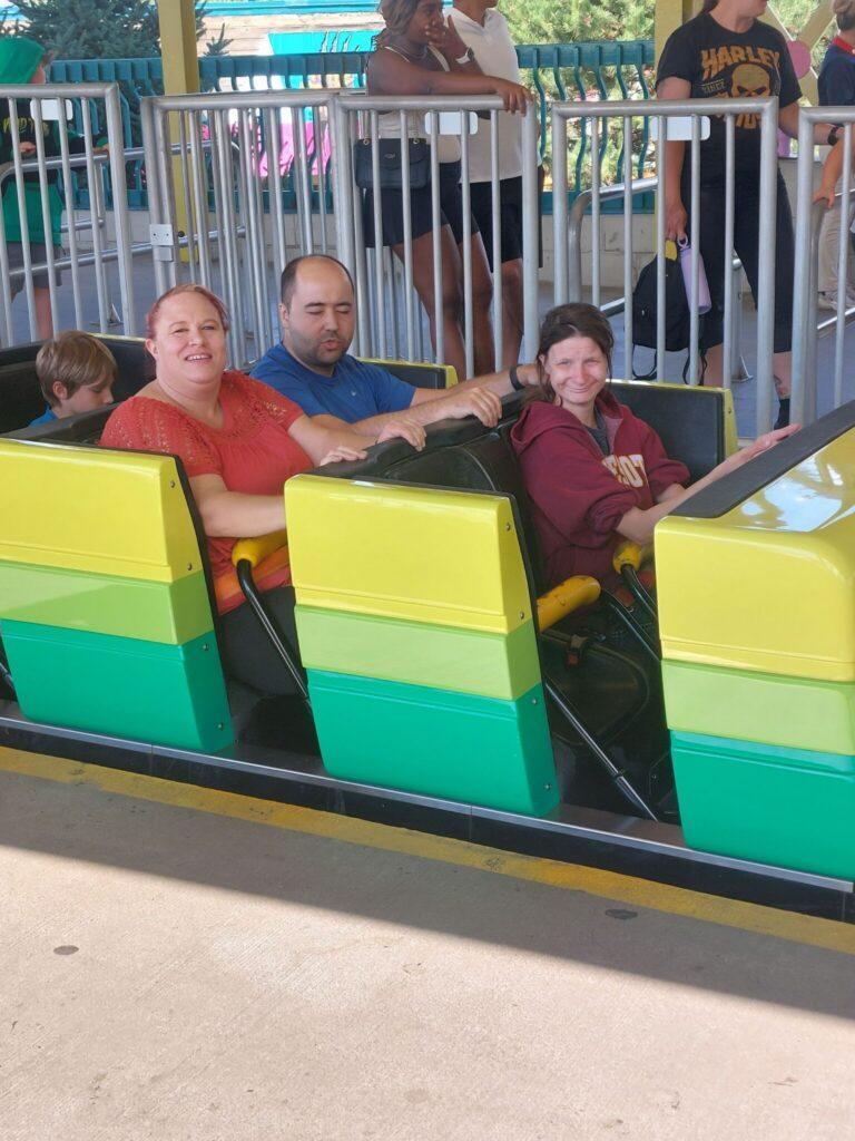 Three people sit in a bright yellow-and-green amusement ride car at an outdoor park on a sunny day.