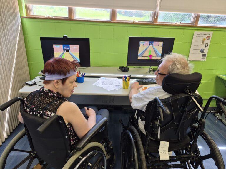 Two people in wheelchairs sit at a table with two computer screens in a bright green room, engaged in an activity.