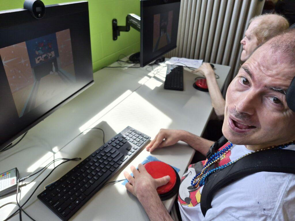 A man with a backpack sits at a desk with two computer monitors and a keyboard, looking at the camera.
