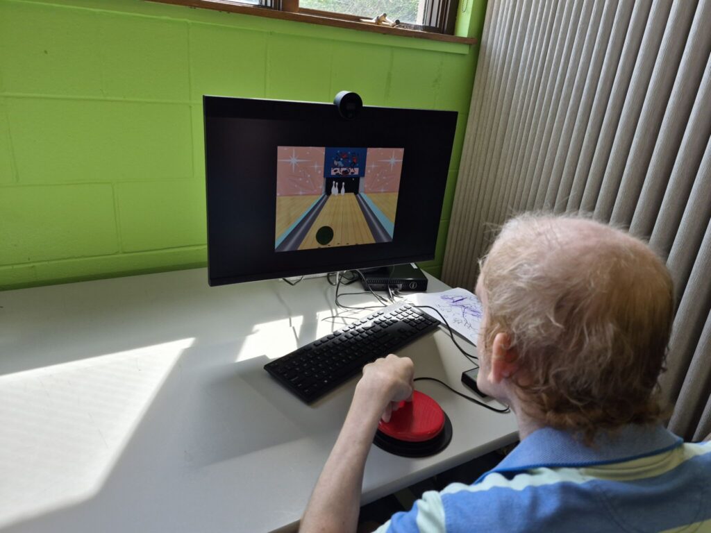 An older man sits at a desk playing a computer game on a small monitor, using a keyboard and a red trackball, in a room with a bright green wall.