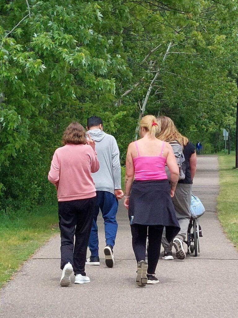 A group of people walking along a paved path in a green park, viewed from behind, with a stroller.