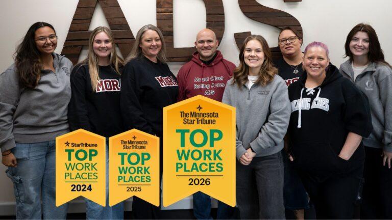 Group of colleagues posing indoors with yellow Top Work Places banners for 2024, 2025 and 2026.