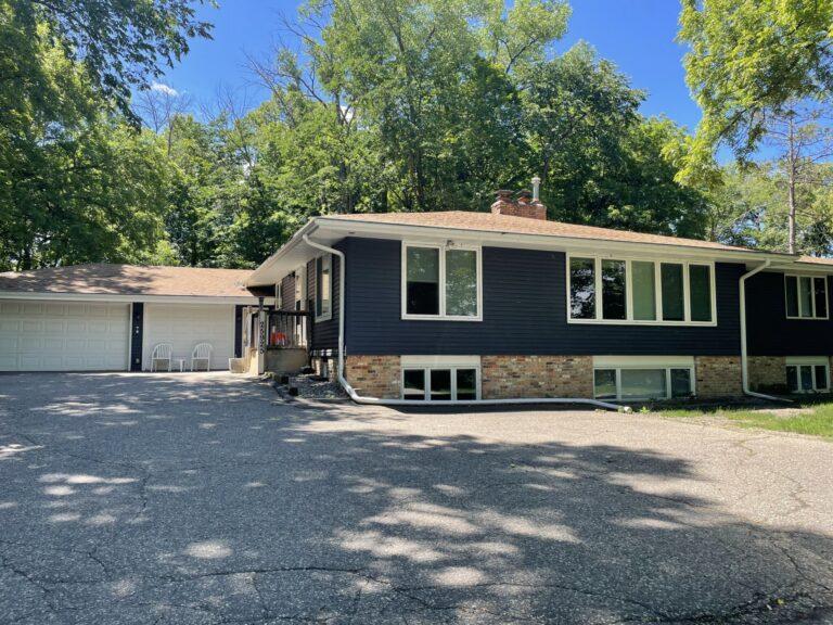A two-story house with dark siding, a stone foundation, and an attached two-car garage, set among trees with a gravel driveway.