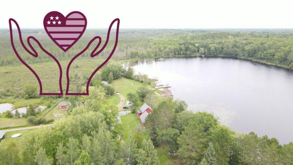 Aerial view of a lake surrounded by dense woods with a few houses along the shore, and a purple hands-holding-heart emblem in the upper left.