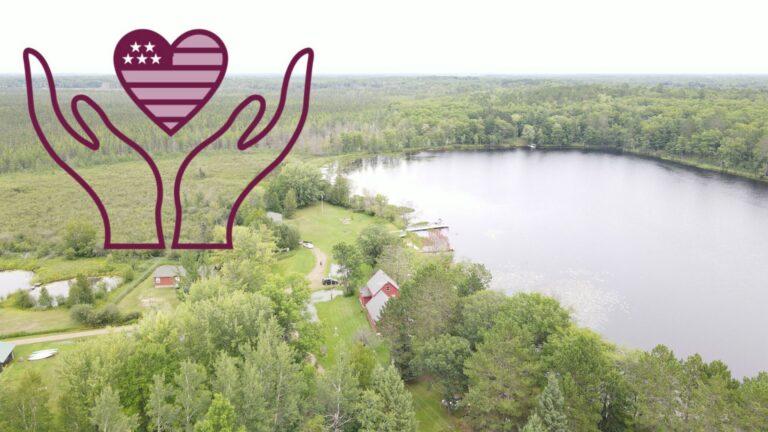 Aerial view of a lake surrounded by dense woods with a few houses along the shore, and a purple hands-holding-heart emblem in the upper left.