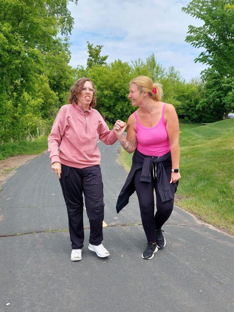Two women walk together along a paved park path, smiling and chatting.