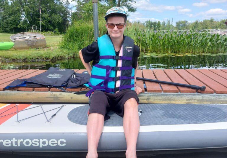 Person wearing a blue life jacket sits on a stand-up paddleboard at a lakeside dock, with a paddle resting on the board.