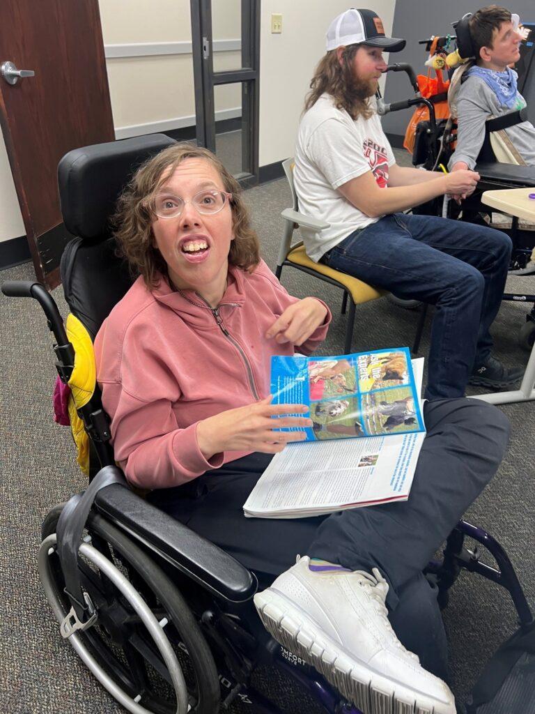 A person in a pink hoodie sits in a wheelchair, smiling while holding a colorful booklet in a classroom.