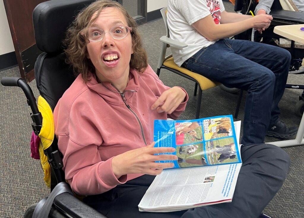 Woman in a wheelchair wearing a pink hoodie sticks her tongue out playfully while holding an open colorful magazine and a newspaper on her lap.