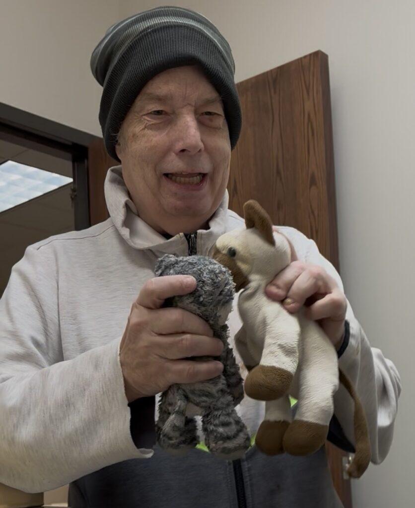 An elderly man wearing a beanie and light jacket smiles while holding two plush toys—a gray shaggy animal and a white‑and‑brown stuffed animal.