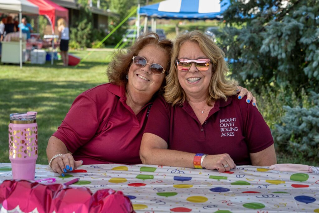 Two smiling women in matching maroon shirts sit at a table outdoors with a colorful tablecloth and a pink tumbler nearby.