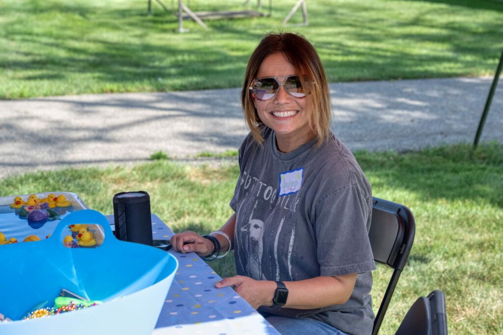 A smiling woman wearing sunglasses sits at an outdoor table with snacks in a park.