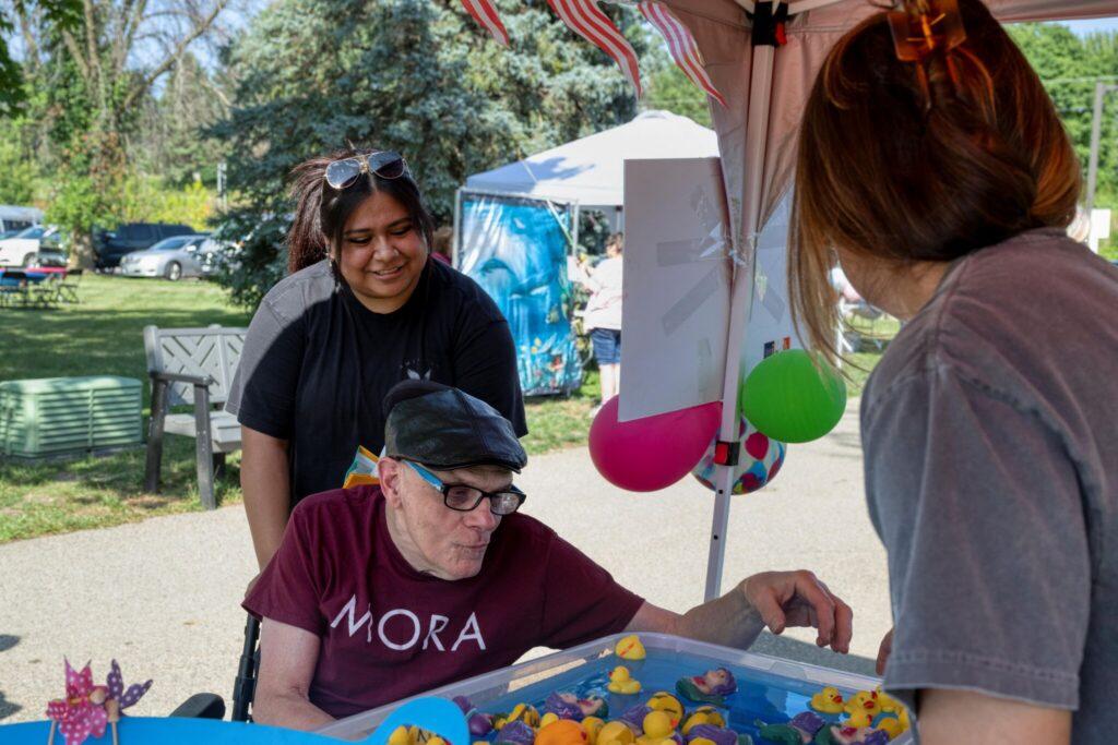 Three people are at an outdoor game booth with a blue tray of yellow prizes, a man wearing a cap at the table, a woman in gray leaning over, and another woman standing behind under a tent with balloons.