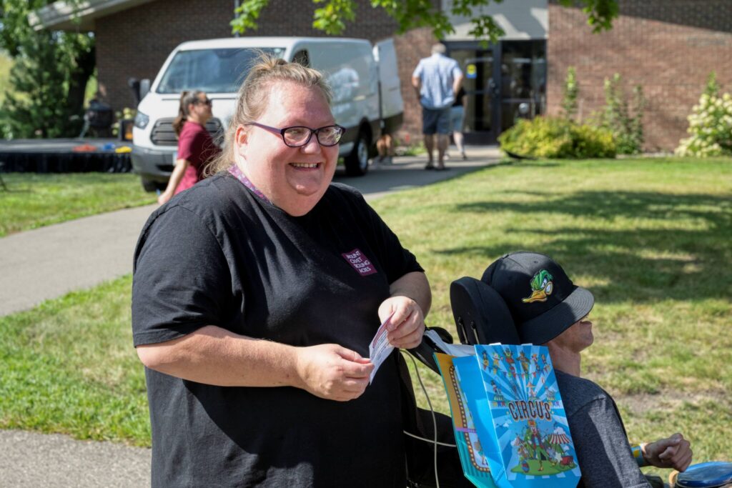 Smiling woman in a black T-shirt hands something to a person in a wheelchair at an outdoor community event.