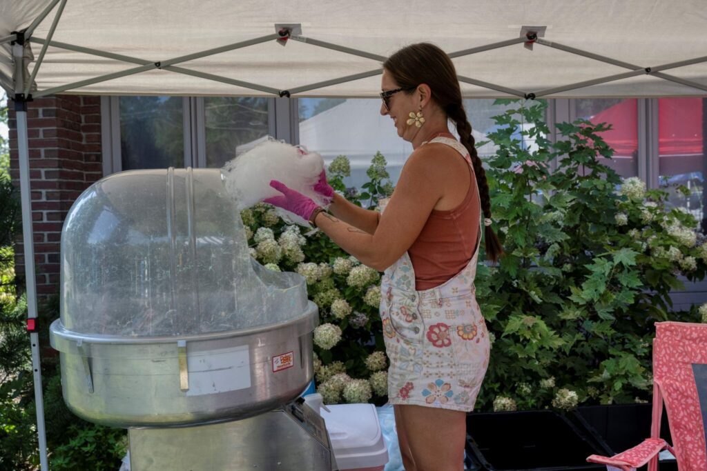 A woman in a floral dress under a canopy prepares pink cotton candy at an outdoor booth.