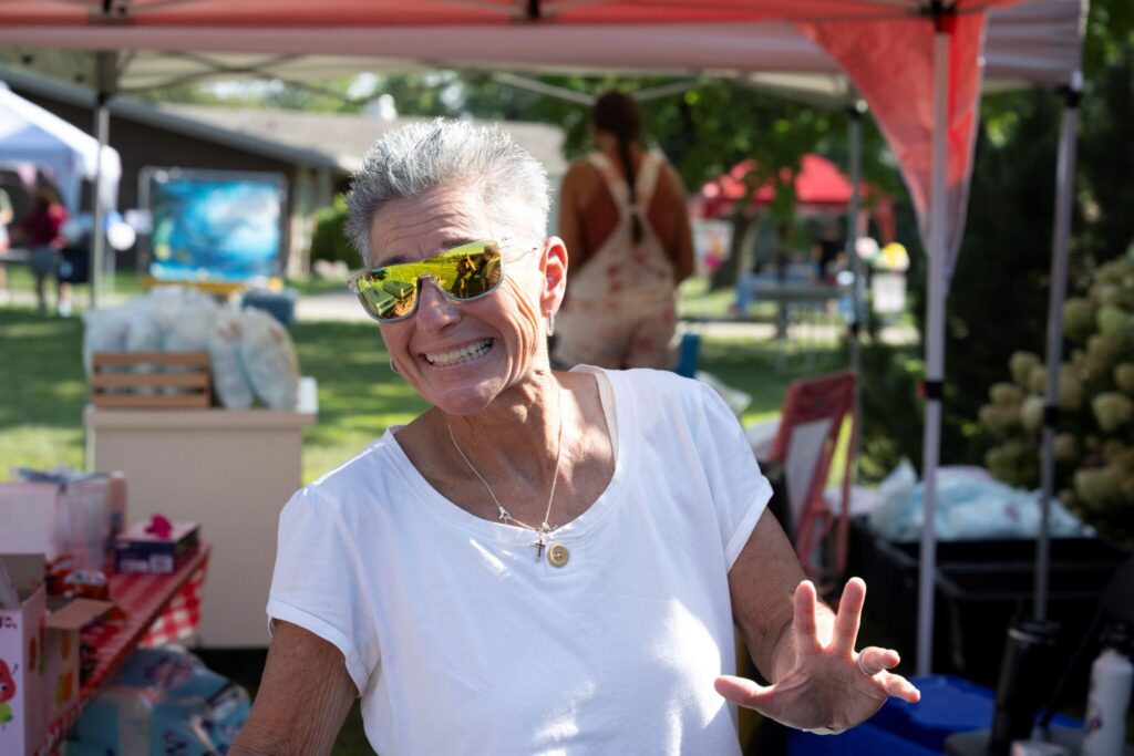 Smiling woman in a white t-shirt and yellow sunglasses waves at an outdoor market under a red canopy.