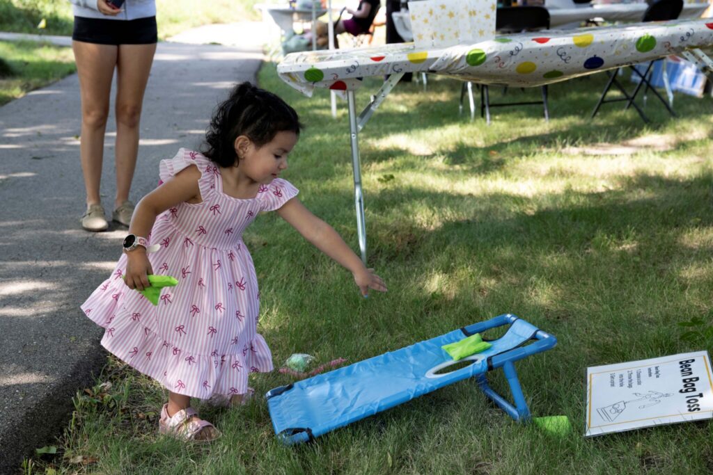 A young girl in a pink dress reaches toward a blue toy airplane on the grass at a park.