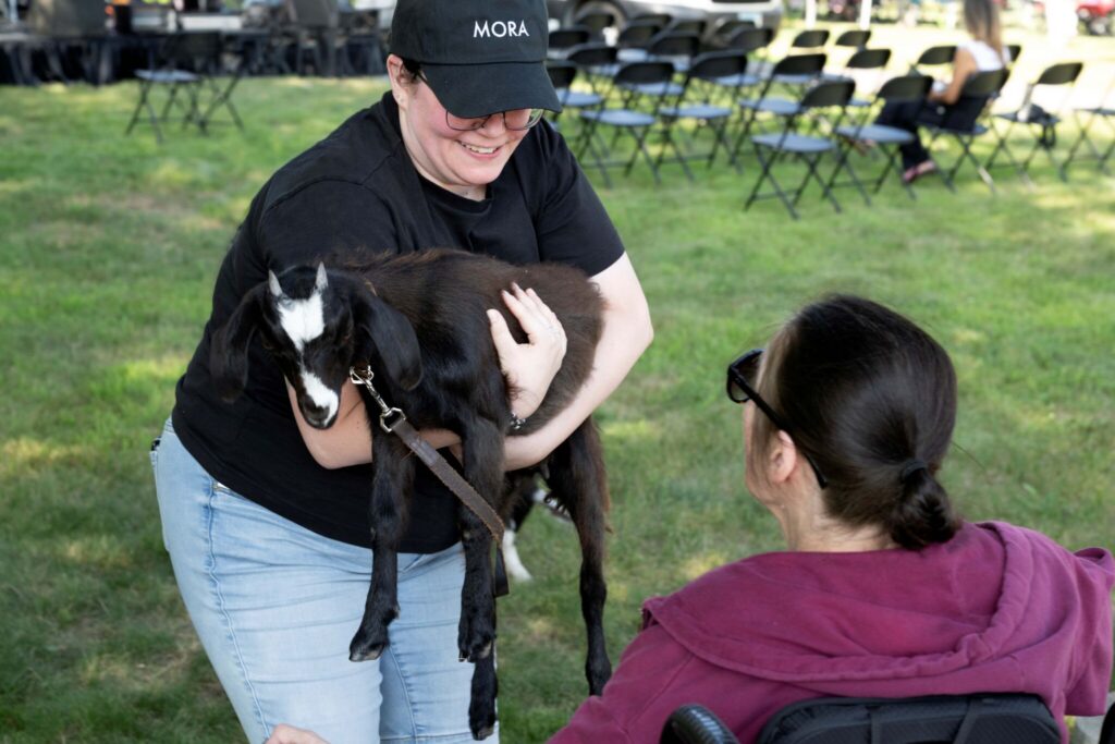 A person wearing a black shirt and cap holds a small brown and black goat while a person in a purple hoodie sits in a wheelchair nearby in a grassy park.