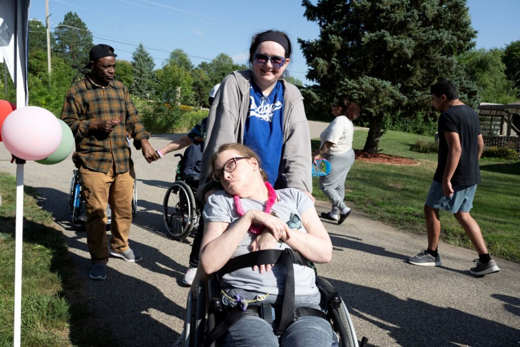 A smiling woman in a wheelchair holds a young child on her lap in a sunny park, with balloons and other people walking in the background.