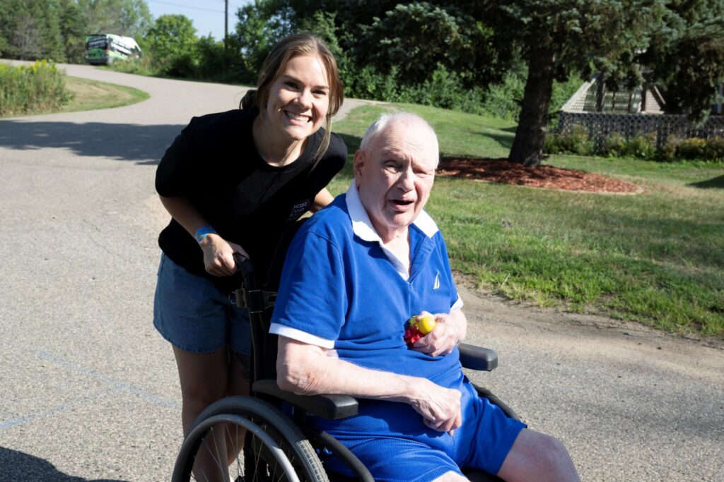 A smiling woman pushes an elderly man in a wheelchair along a sunny suburban street.