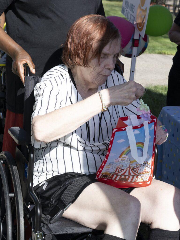 An elderly woman sitting in a wheelchair opening a red gift bag outdoors.