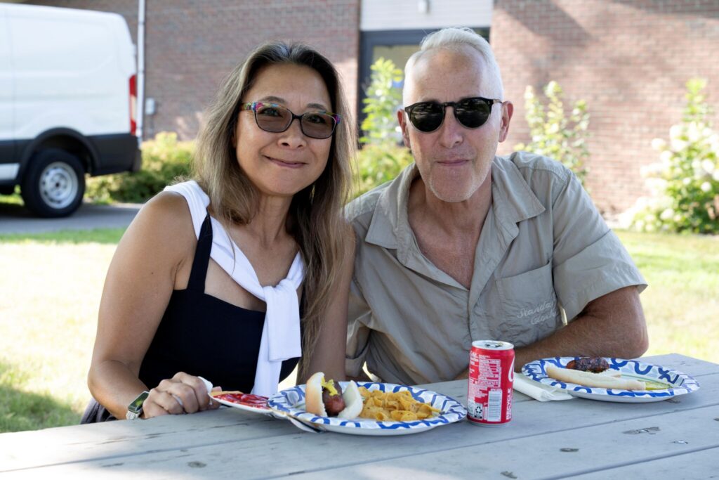 A man and a woman sit at a picnic table outdoors, eating from paper plates with drinks.