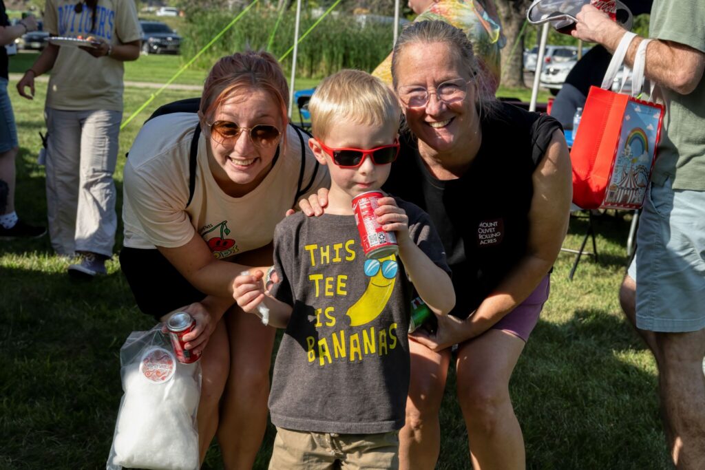 Two smiling women kneel beside a boy wearing sunglasses at a sunny outdoor park.