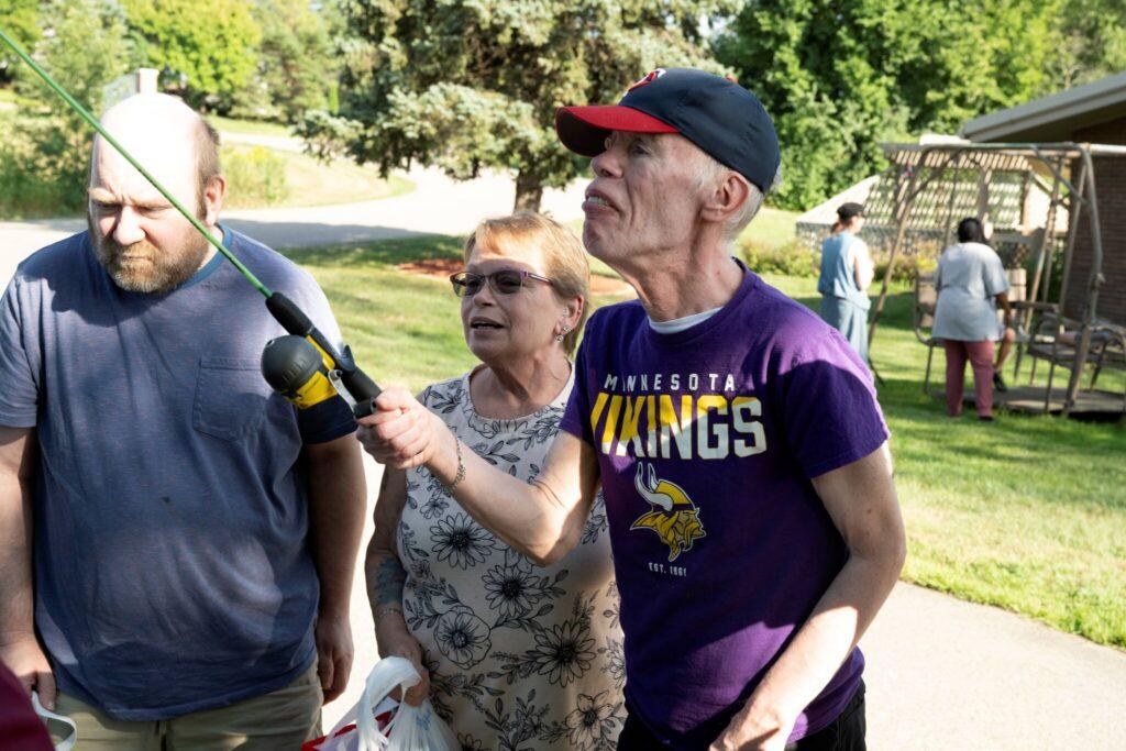 Three people stand outdoors in a park on a sunny day, with a man in a purple Vikings shirt holding a microphone toward the others.