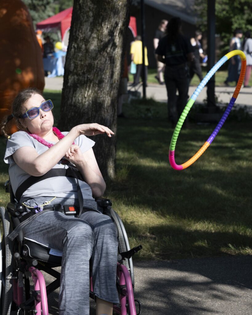 A person in a wheelchair outdoors in a park, wearing sunglasses, with a colorful hula hoop spinning nearby.