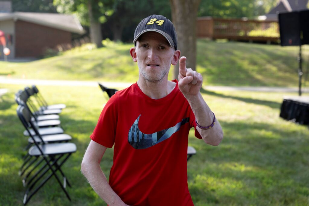 A man in a red shirt and black cap stands on a grassy lawn, raising one finger, with folding chairs in the background.