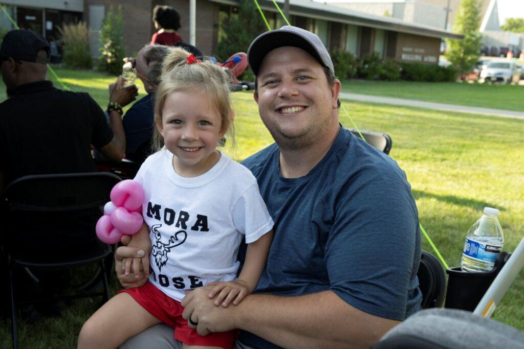 A man in a blue T-shirt and cap sits outdoors with a smiling young girl in a white T-shirt, the girl holding a pink plush toy.