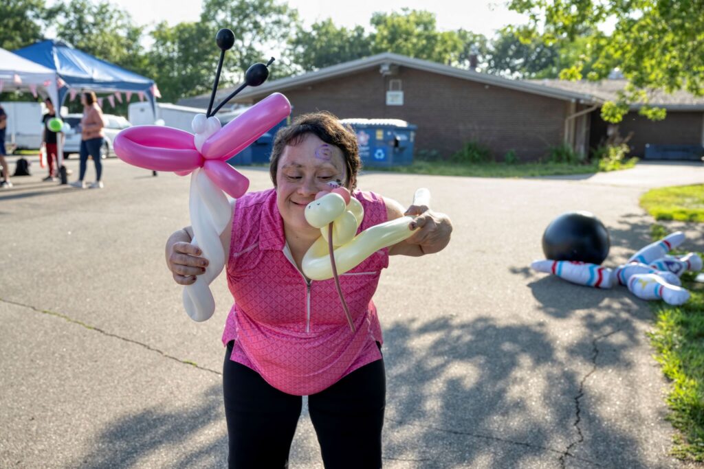 Smiling woman in a pink shirt holds two oversized balloon animals outdoors.