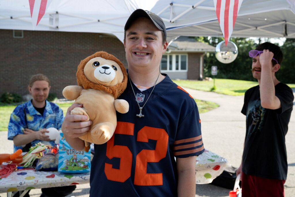 Smiling man in a blue jersey with the number 52 holds a teddy bear at an outdoor booth.