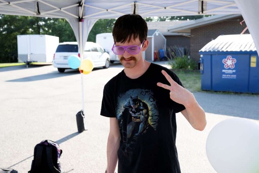 A man with a mustache wearing a black graphic t-shirt stands under a white canopy outdoors, making a peace sign.